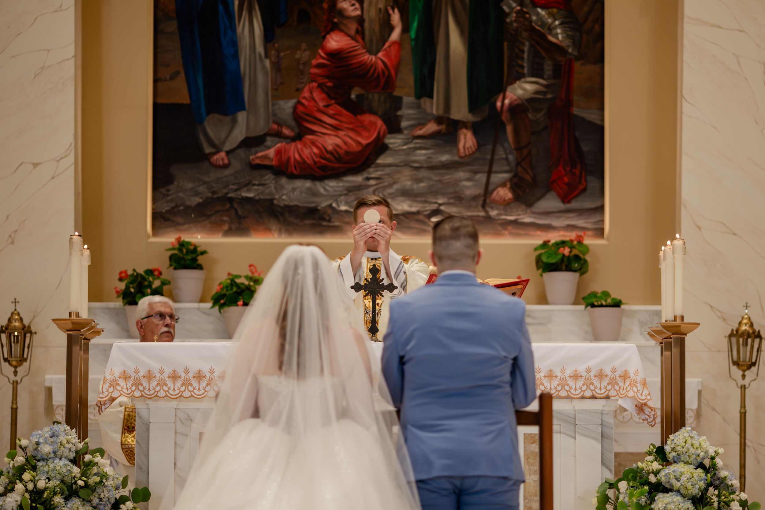 Wedding guests stand in a grand reception hall decorated with white flowers and elegant candelabras.