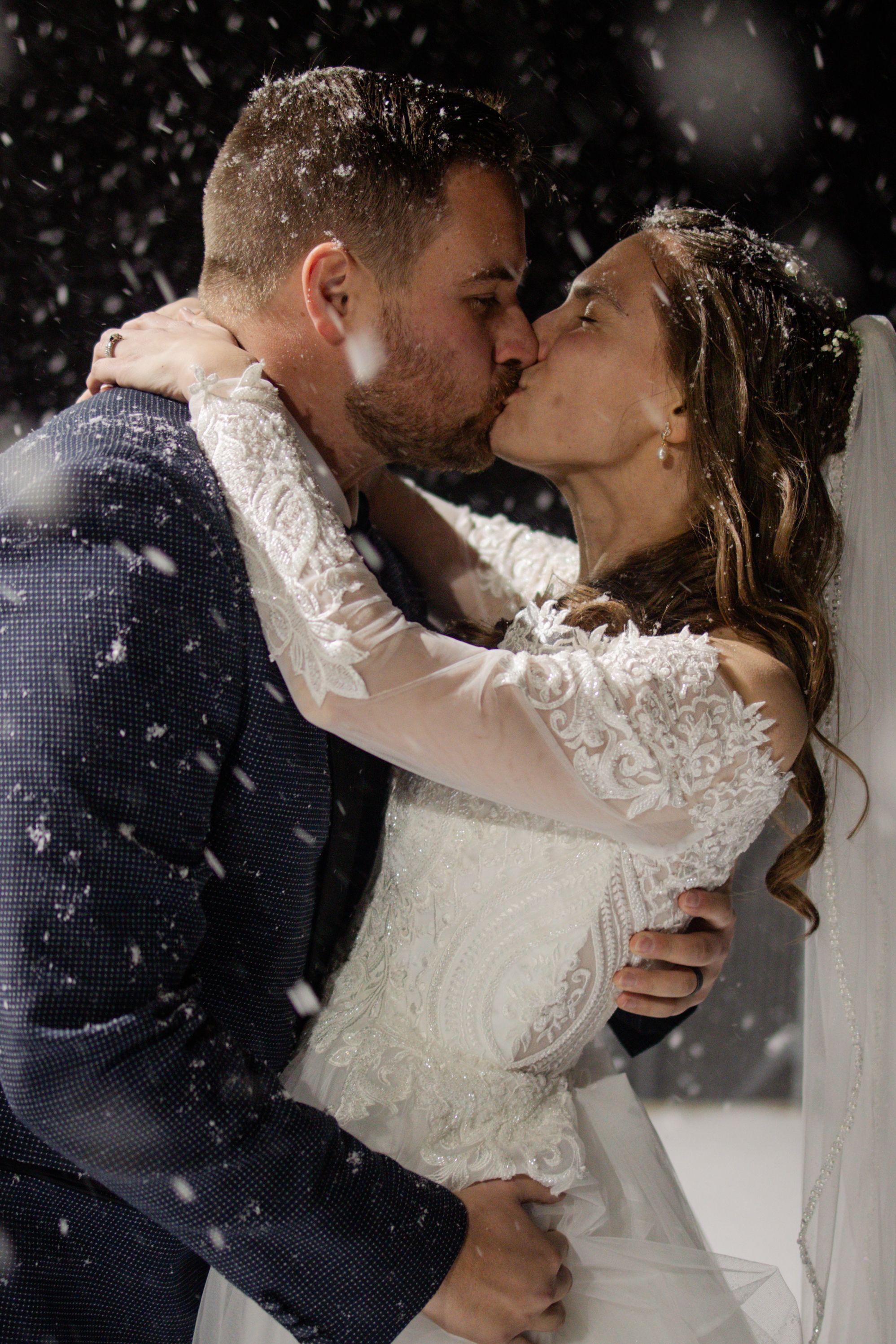 A romantic winter wedding scene with a couple kissing in falling snow, the bride wearing a long-sleeved lace gown.