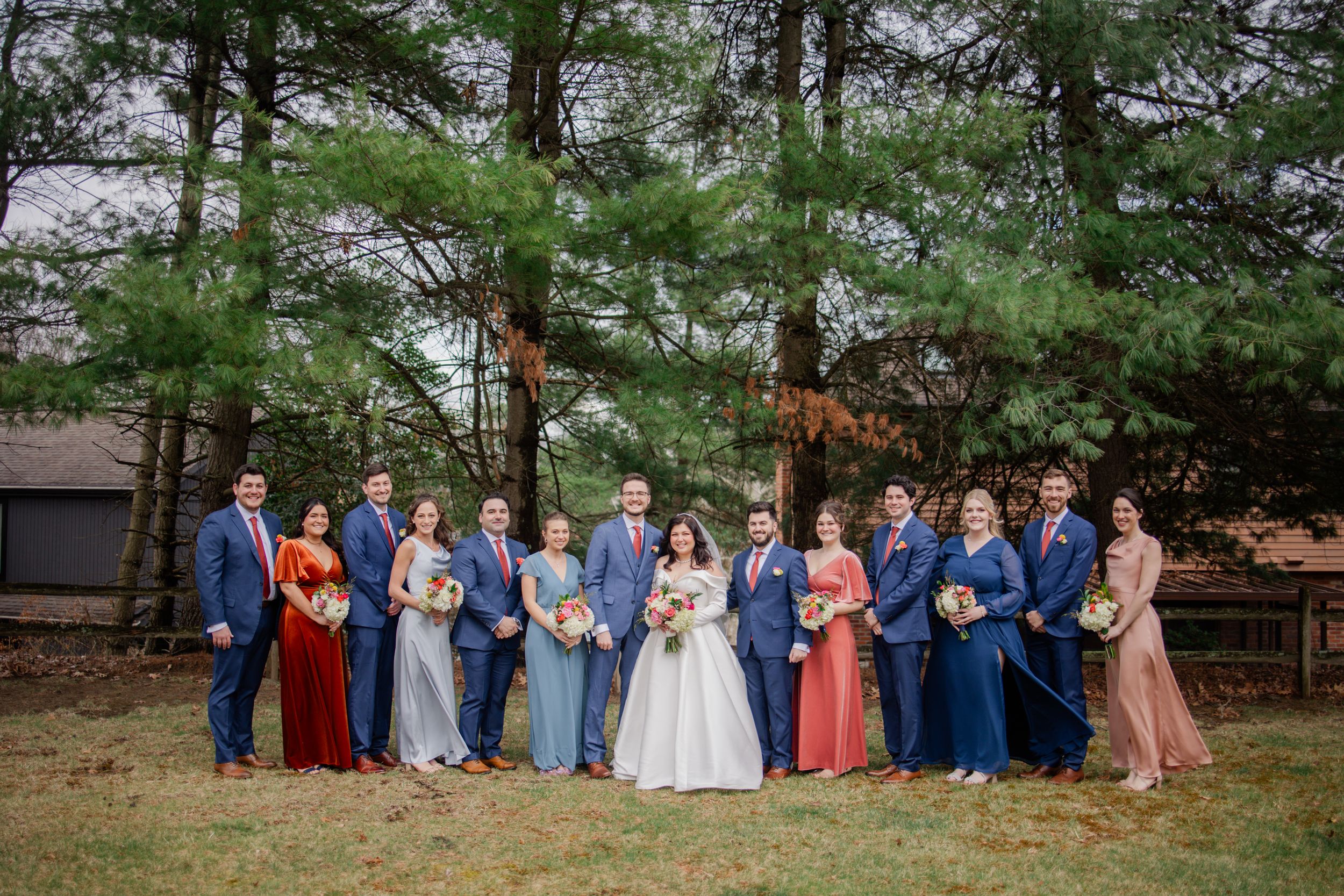 Wedding party poses in front of evergreen trees wearing navy blue and coral colored formal attire.