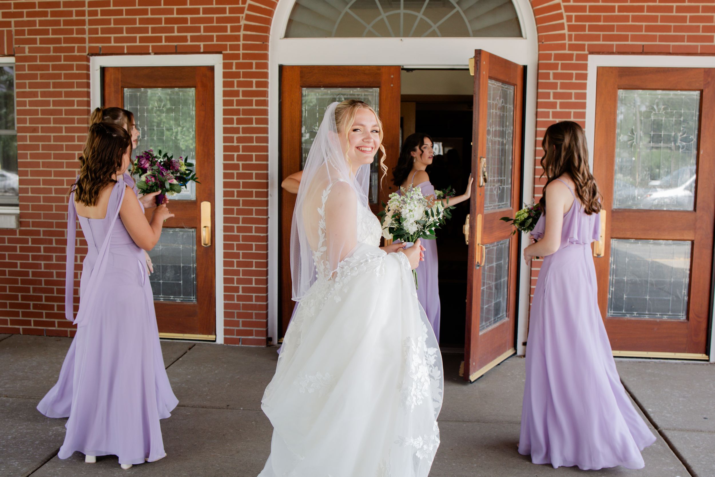 Bridesmaids in lavender dresses stand with a bride in white along a brick building's covered walkway.