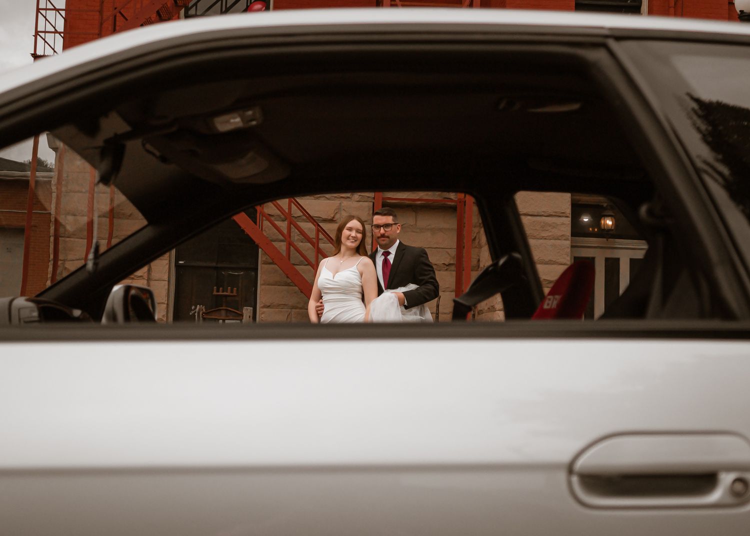 Reflection of a wedding couple in a car window with brick building in background.