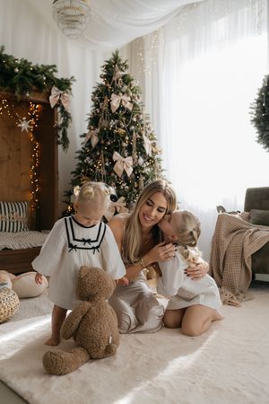 Smiling mother kisses her daughter as a toddler stands by a teddy bear in front of a decorated Christmas tree.