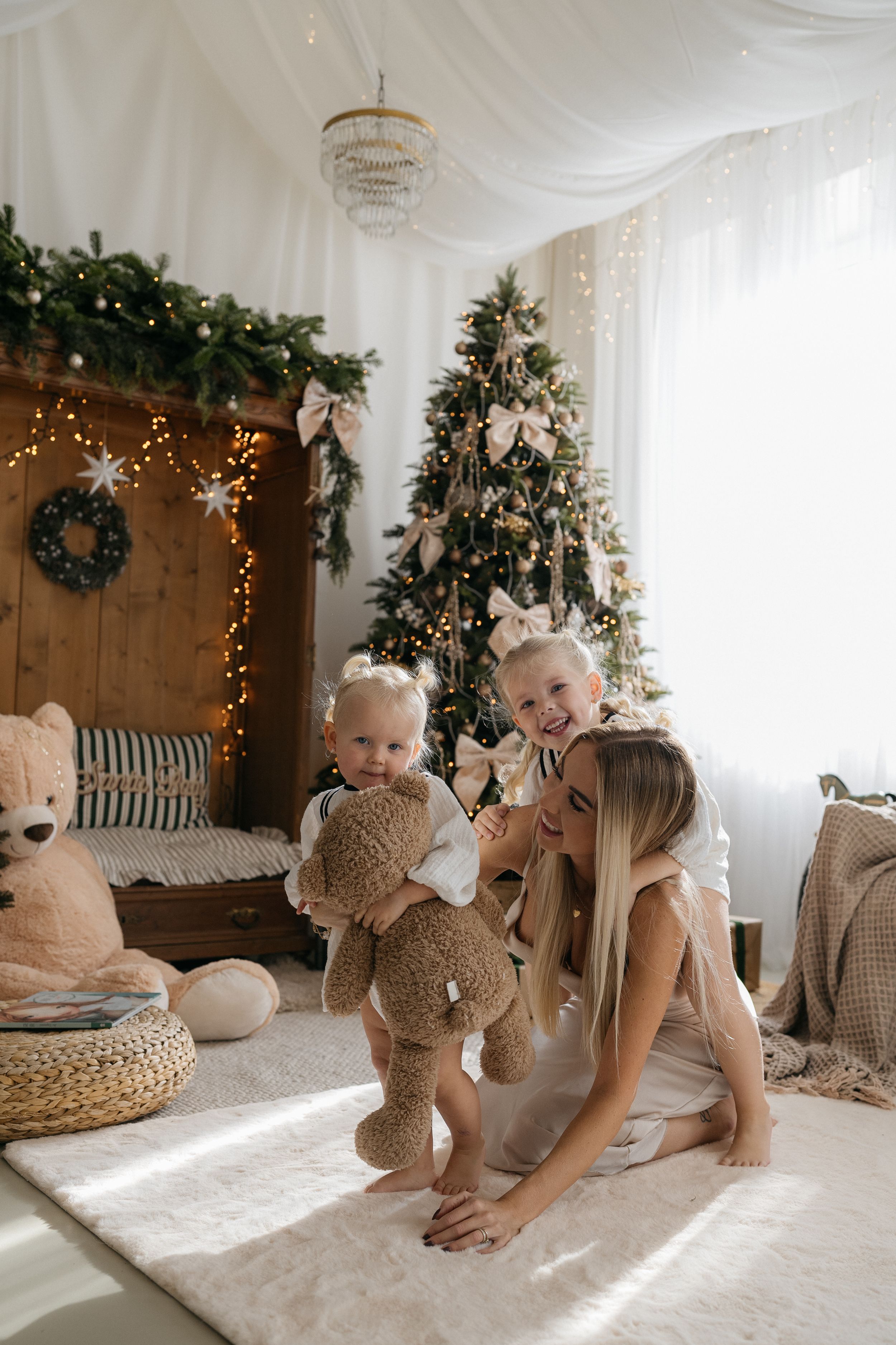 Mother plays on a white rug with two young children holding teddy bears in a festive room with a Christmas tree.