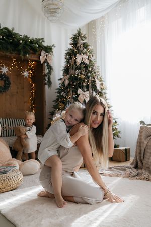 Mother sits on a fluffy rug as her daughter hugs her back, with a toddler and a decorated Christmas tree behind them.