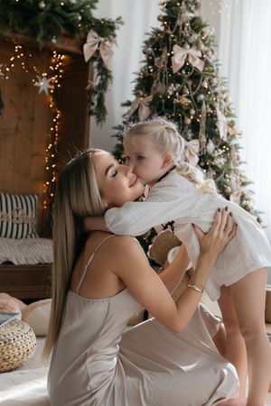 Mother in a satin dress holds her little girl in a tender cheek kiss beside a Christmas tree with twinkling lights.