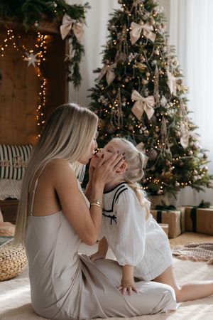Mother in a silk dress gently kisses her daughter's face while sitting by a glowing Christmas tree and wrapped presents.