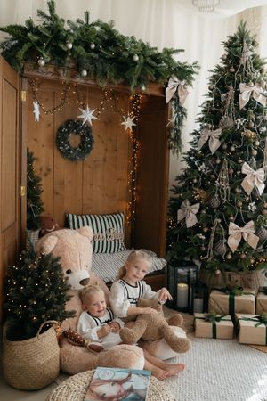 Two young girls sit with a large teddy bear beside a Christmas tree, gifts, candle lantern and a rustic wooden alcove.