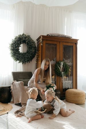 Mother hands a gift to two small children playing on a cozy rug near a Christmas wreath and a vintage wooden cabinet.