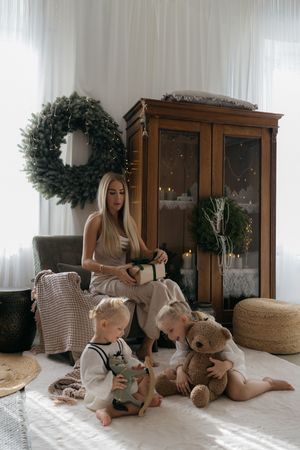 A woman holding a wrapped gift sits by a Christmas wreath and wooden cabinet while two toddlers play with toys beside her.