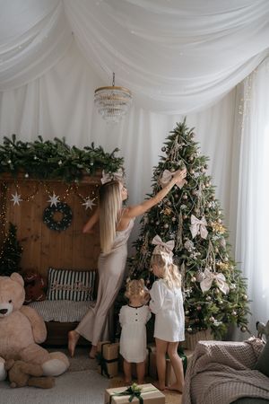 A woman reaches to decorate a Christmas tree with bows while two little girls stand nearby beside gifts and a large teddy bear.