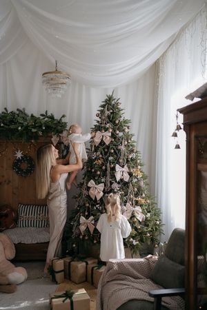 A woman lifts a toddler toward a bow-decorated Christmas tree while a young girl in white watches, with wrapped gifts below.