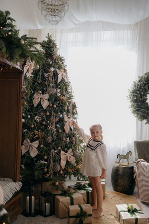 A little girl in a white dress touches a Christmas tree decorated with bows and ornaments, with wrapped presents at her feet.