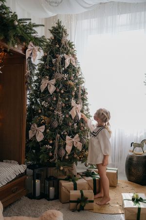 A young girl in a white sailor-style dress reaches toward a beautifully decorated Christmas tree beside gifts and candle lanterns.