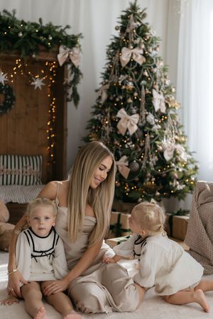 A smiling mother sits on a cozy rug with two little girls in front of a glowing, bow-trimmed Christmas tree.