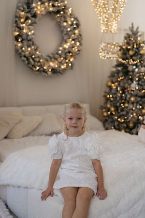 A little girl in a white puff-sleeve dress sits on a white bed with a lit Christmas wreath and gold-ornamented tree behind her.