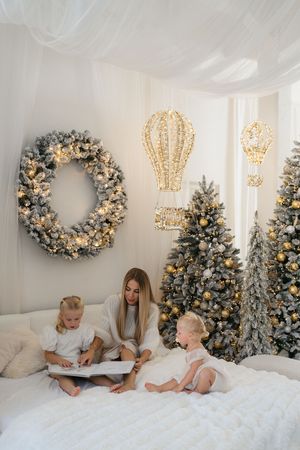 Mother reads a book with two young daughters on a cozy white bed surrounded by Christmas trees, a wreath and glowing balloon lights.