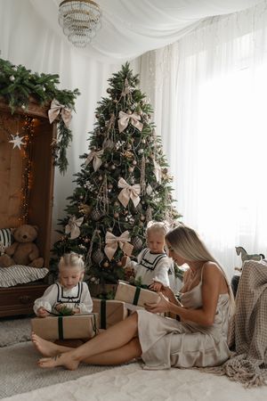 Mother and two children in neutral outfits open wrapped gifts beside a Christmas tree decorated with cream bows and a teddy bear.