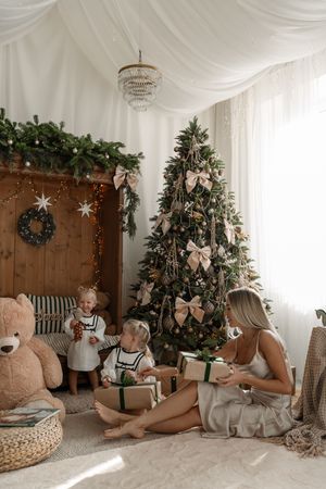 Woman holds a gift while two toddlers sit by a bow-decorated Christmas tree, a giant teddy bear and a wooden mantel with garland.