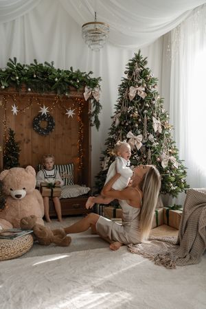 Mother sits on a fluffy rug lifting a smiling baby beside a bow-trimmed Christmas tree, giant teddy bear and a child with a gift.