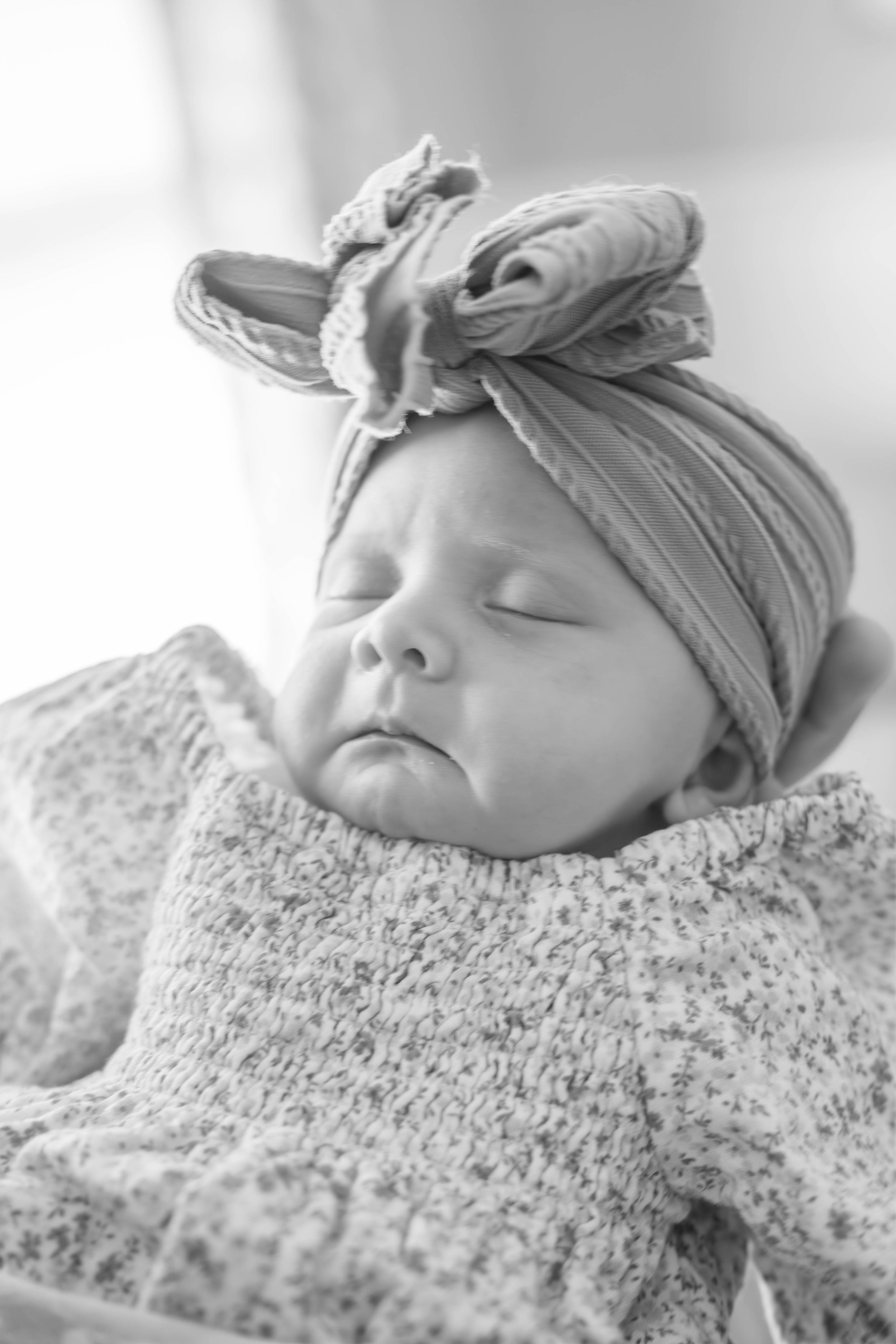 A sleeping newborn baby wearing a gray headband rests peacefully on textured white blanket.