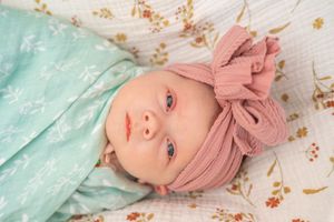 Baby wearing mint green outfit and pink bow headband lies on floral fabric background.