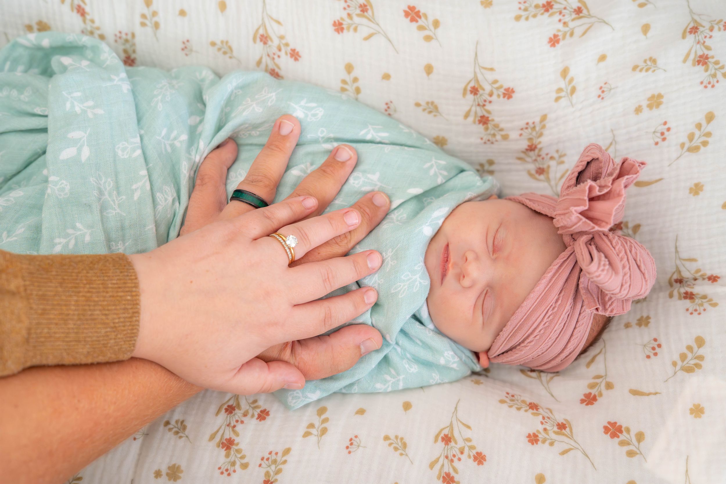 Small hands reach towards baby in mint green outfit lying on floral patterned fabric.