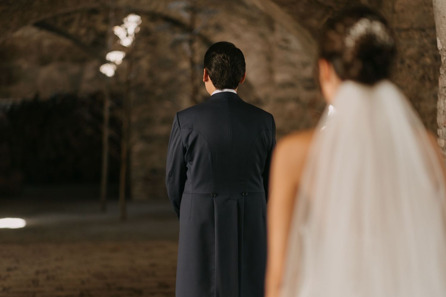 A couple in wedding attire stands together in a dimly lit rustic venue with stone walls.