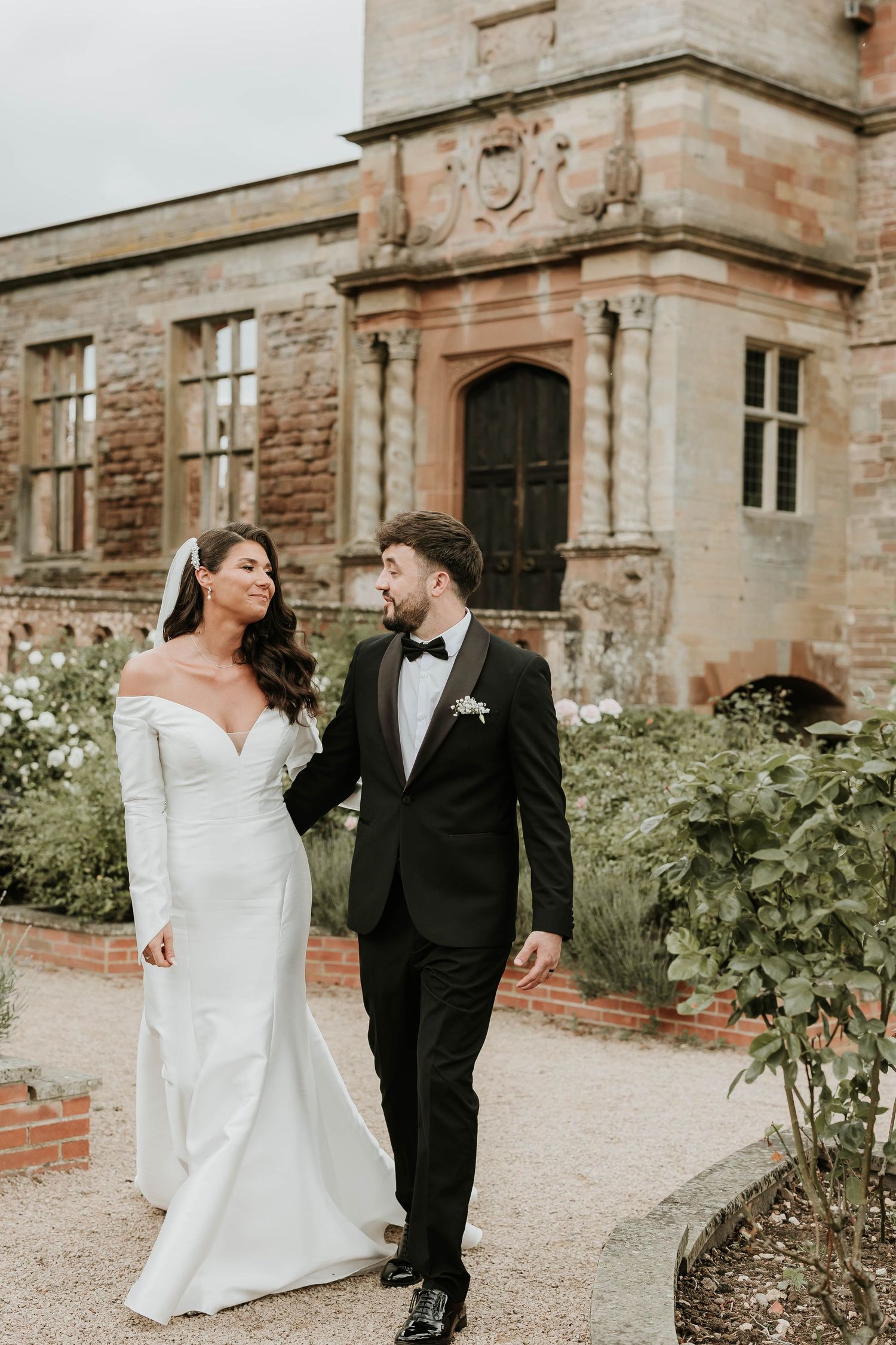 A romantic wedding couple poses together in front of a historic brick building with lush greenery.