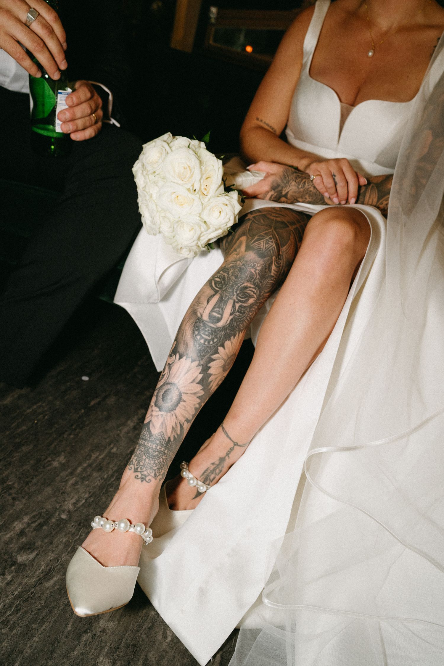 Close-up view of tattooed legs next to white wedding dress fabric on dark wooden floor.