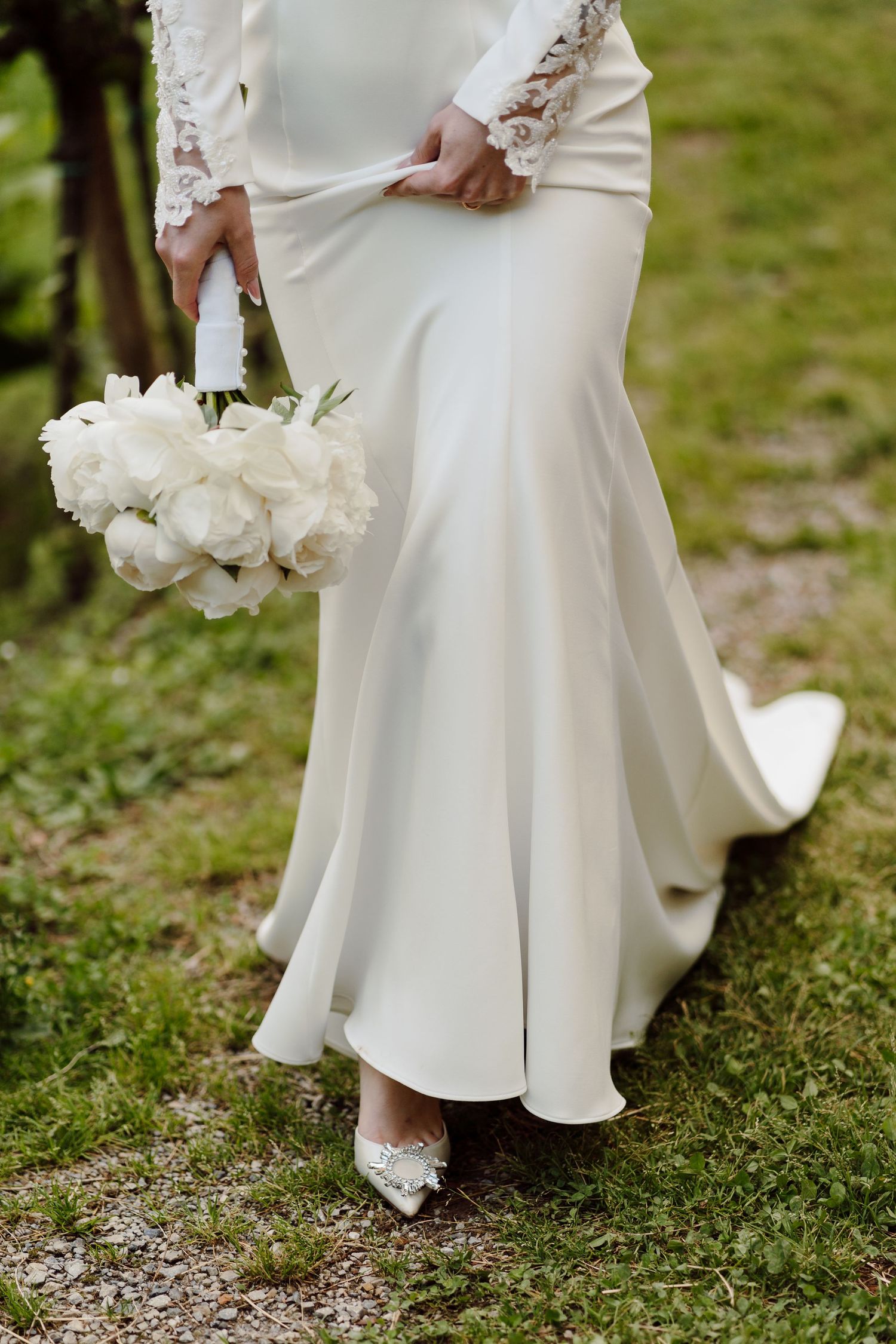 Close-up of bride in white mermaid wedding gown holding white peony bouquet with lace sleeves.