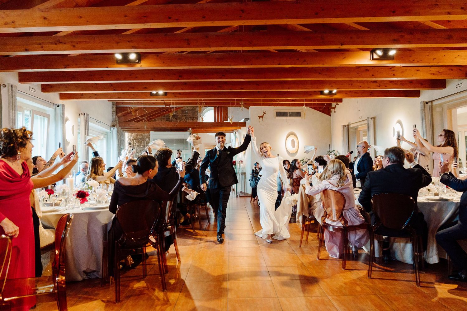 Bride and groom making grand entrance into rustic reception hall as guests cheer and celebrate.