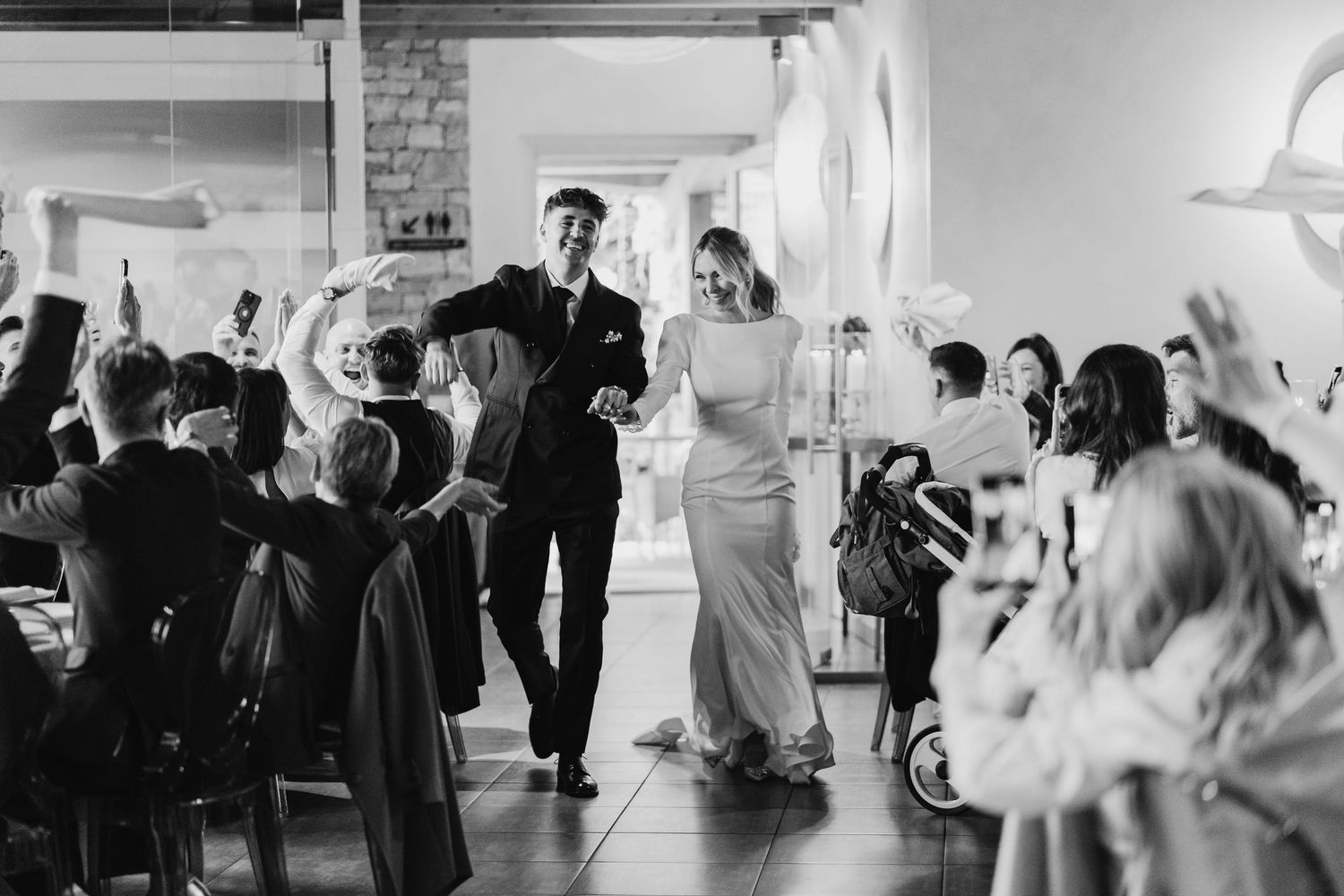Black and white photo of newlyweds walking into wedding reception as guests raise hands in celebration.