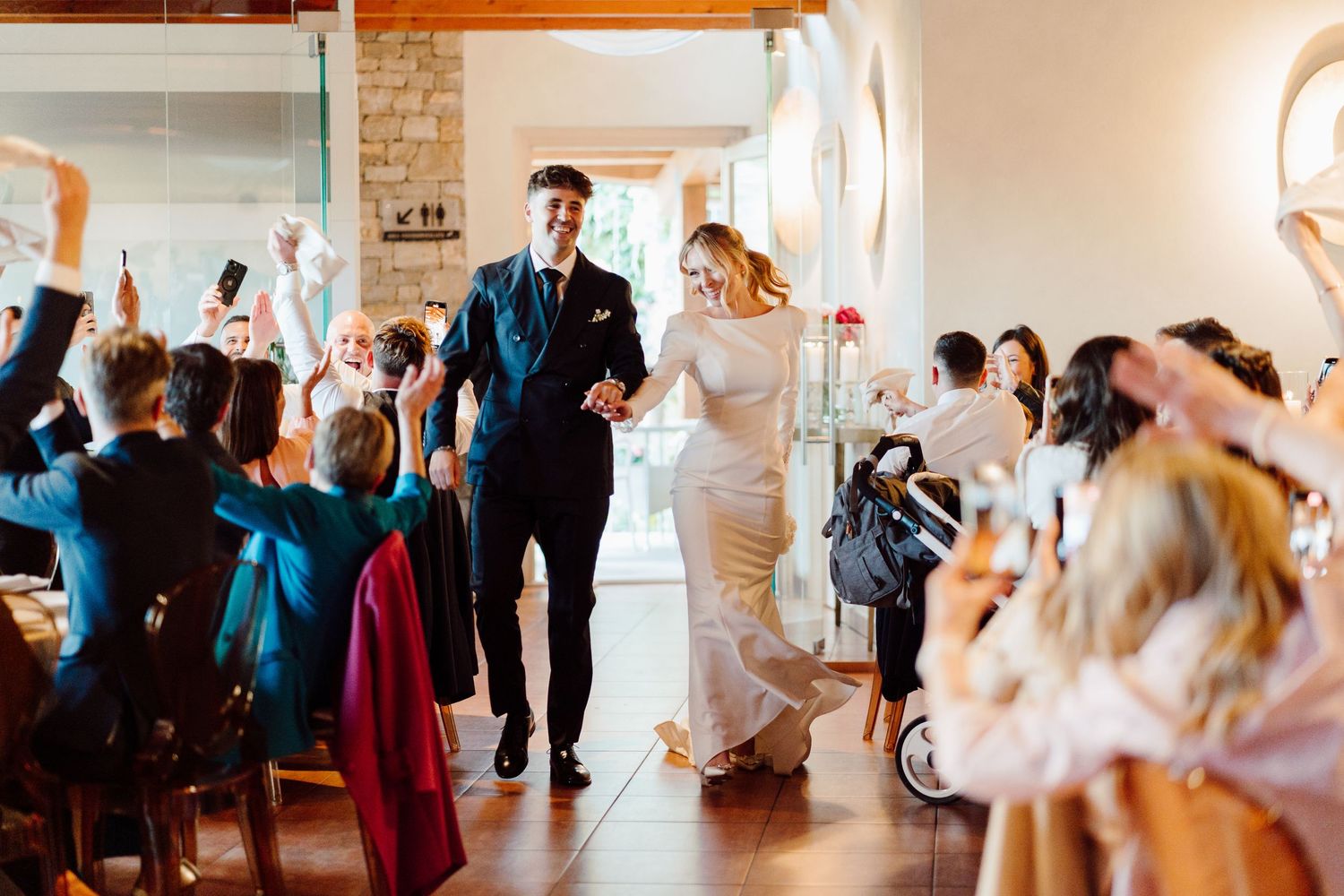 Smiling bride and groom entering elegant wedding reception venue as guests cheer and take photos.