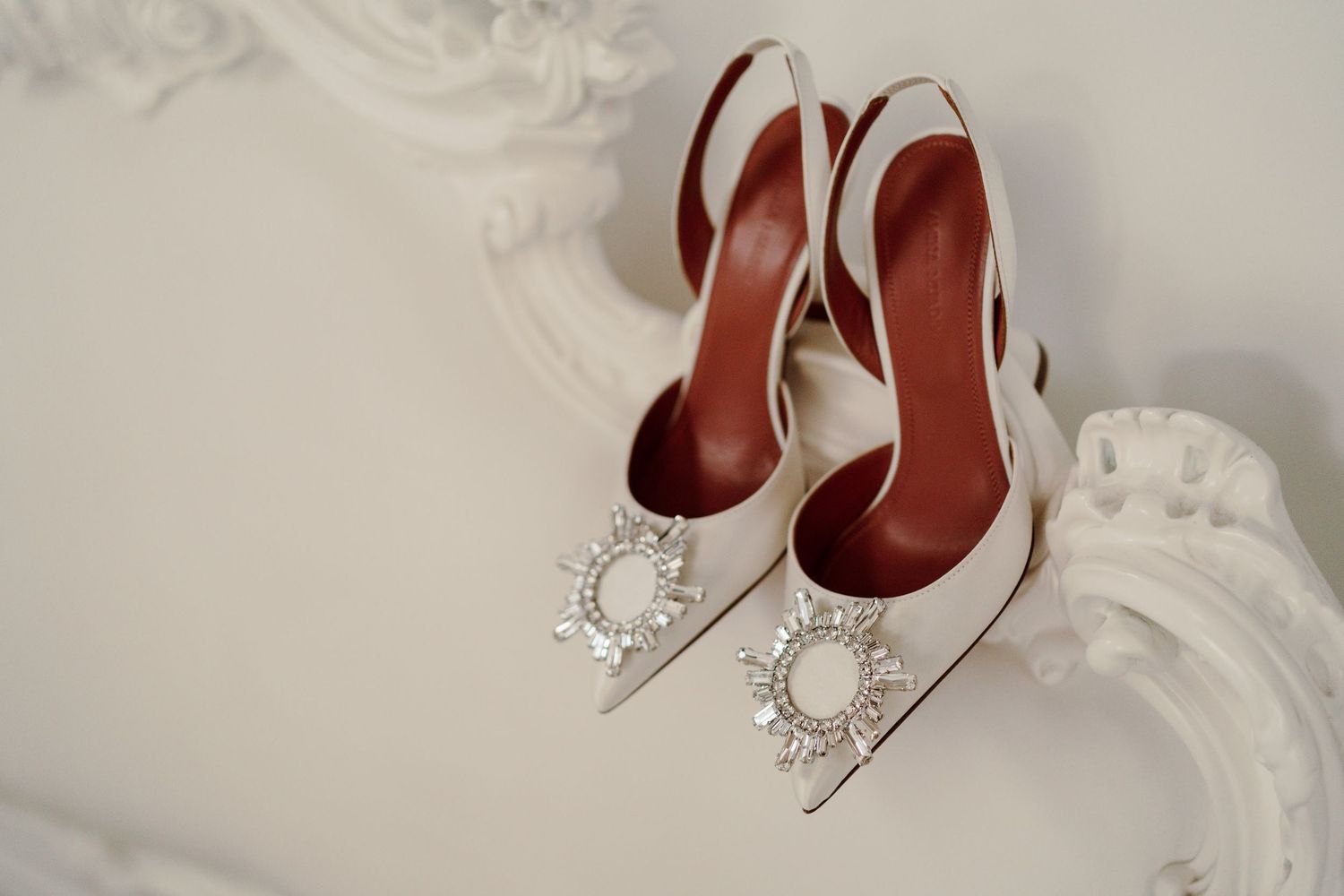 Elegant white bridal heels with crystal embellishments displayed on ornate white frame.