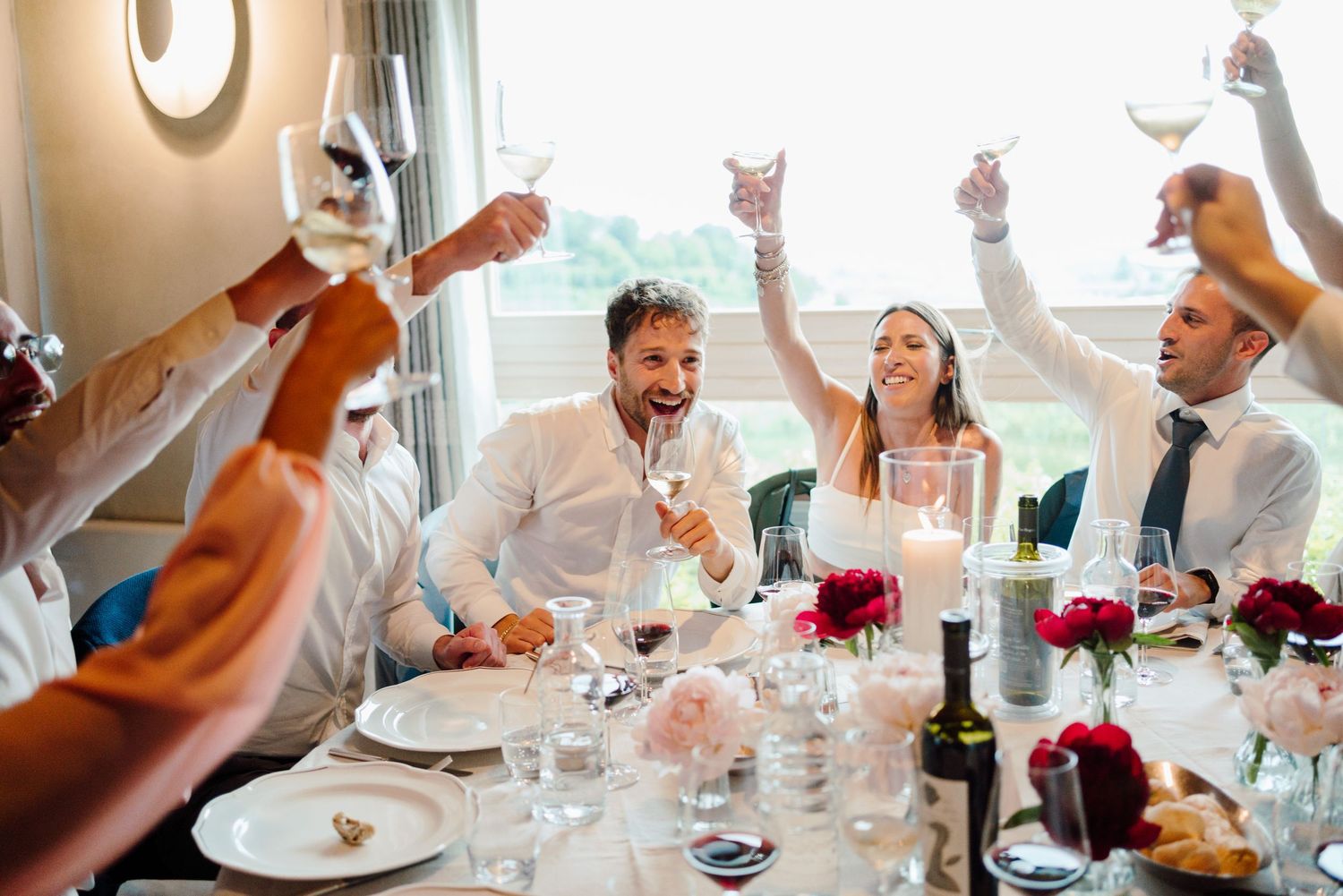 Happy wedding party raising wine glasses in a colorful toast at a floral decorated table.