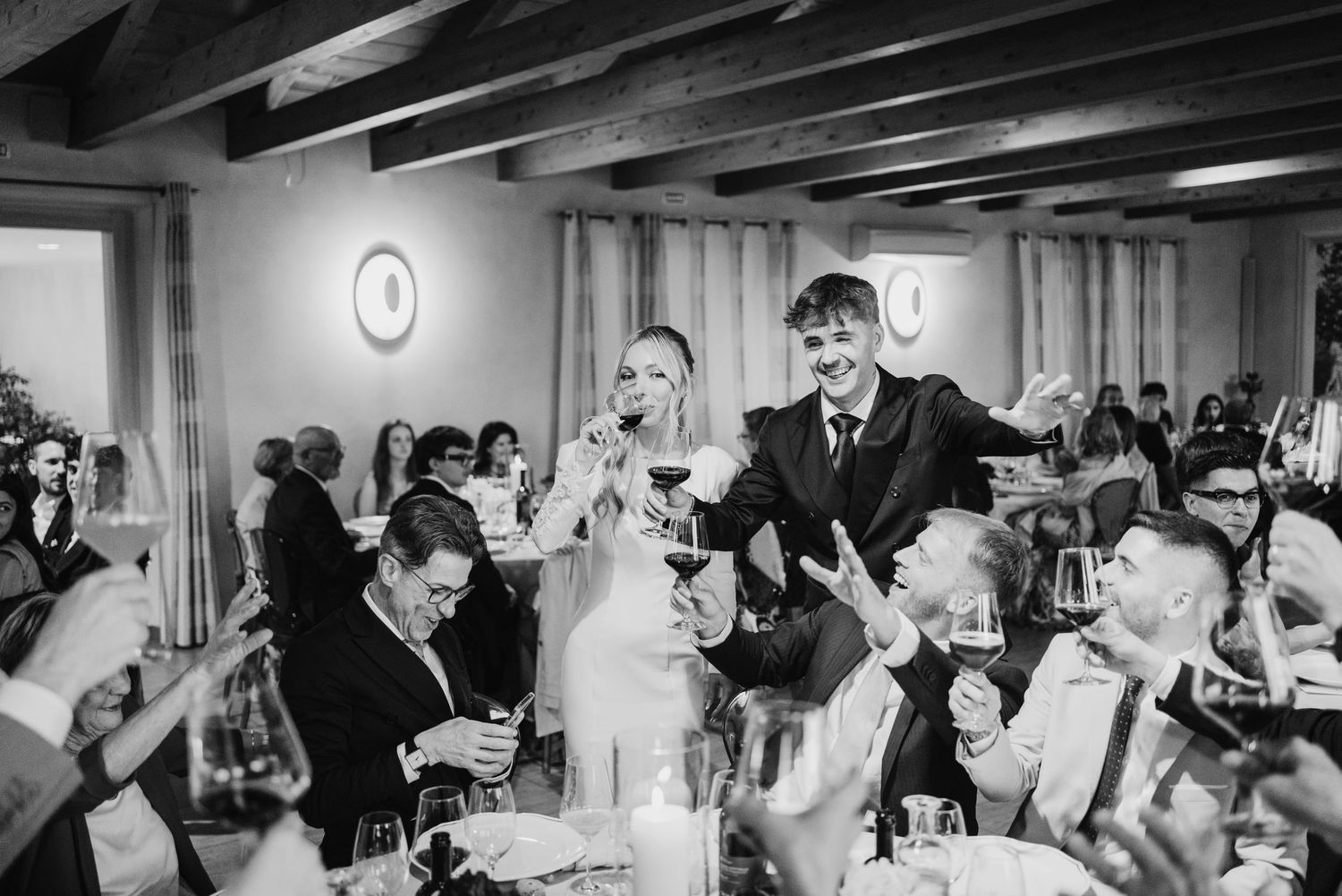Bride and groom standing at their table as guests raise glasses in a lively wedding toast.
