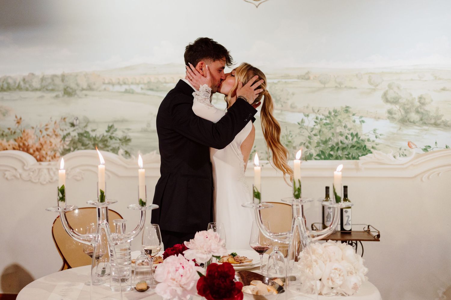 Bride and groom sharing a romantic kiss at their candlelit sweetheart table with floral mural backdrop.