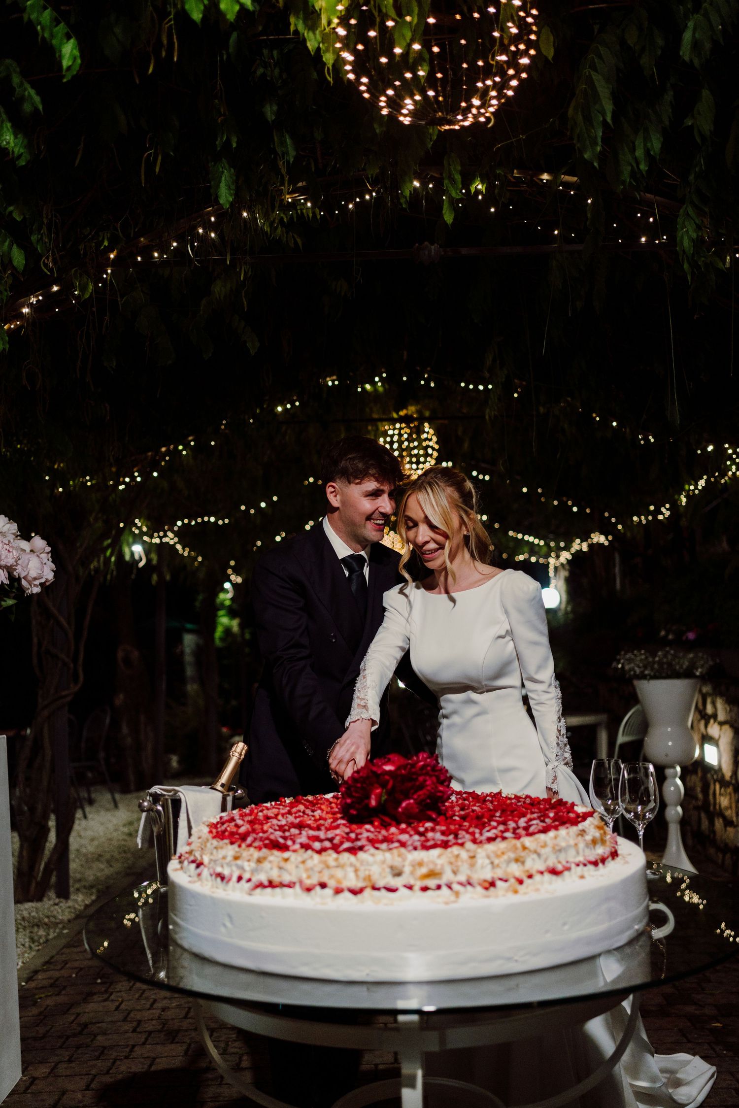 Bride and groom smiling together as they cut a large heart-shaped wedding cake outdoors at night.