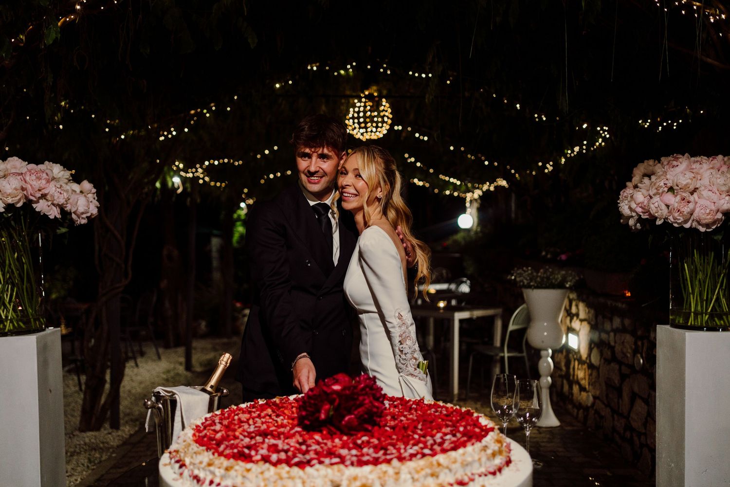 Smiling bride and groom cutting a large round wedding cake decorated with red rose petals at night.