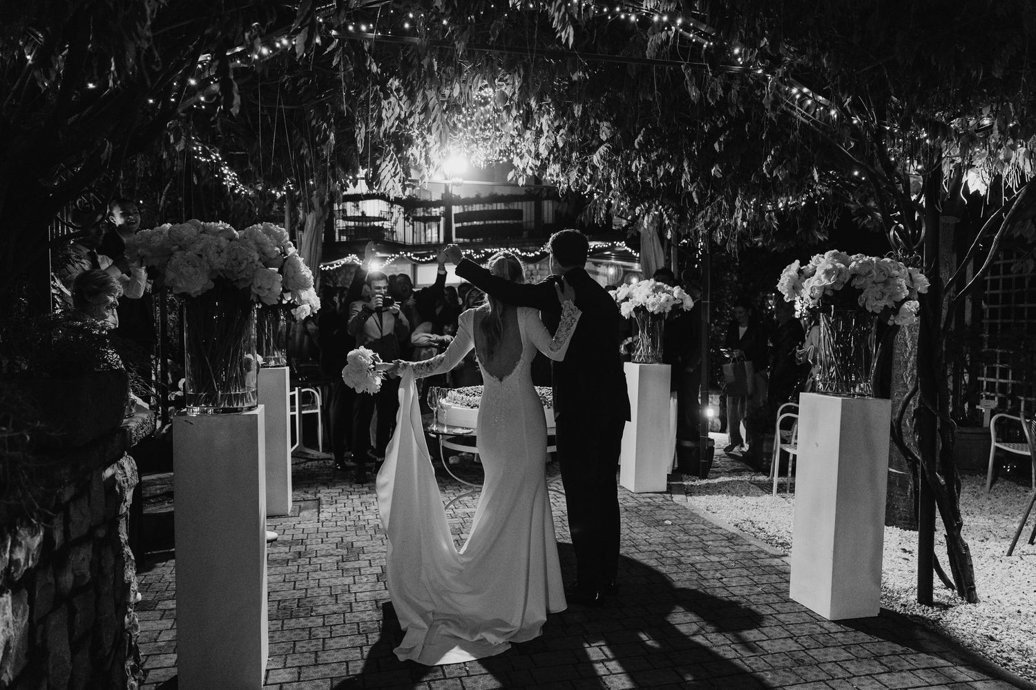 Bride and groom walking through a romantic outdoor aisle lined with white floral arrangements at night.