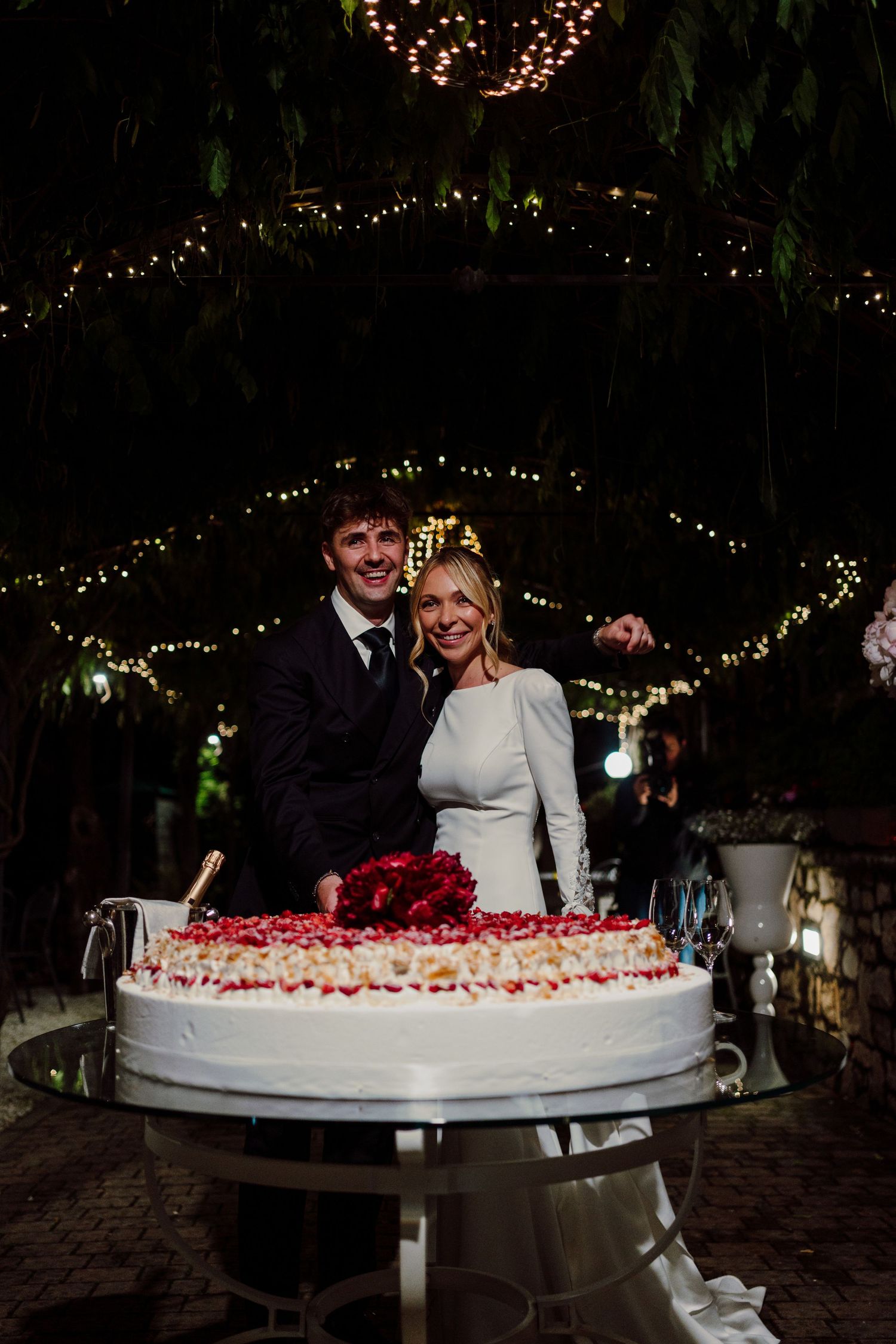 Bride and groom smiling behind elegant white wedding cake decorated with red roses under fairy lights at night.