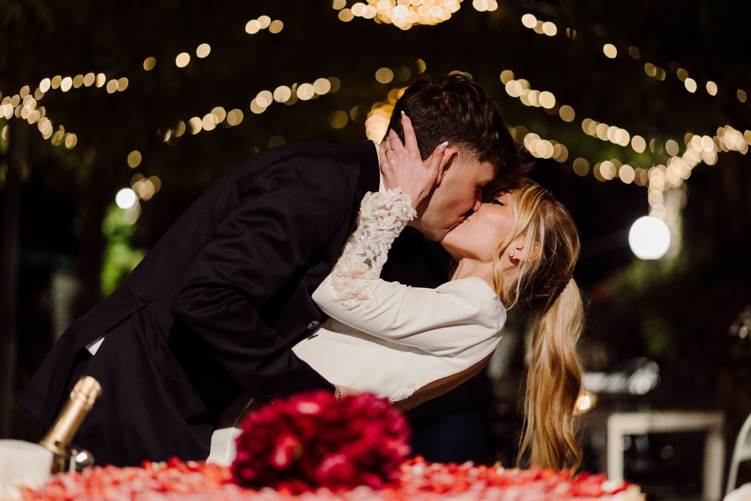 Bride and groom sharing a romantic kiss near red roses and champagne under warm fairy lights at night.
