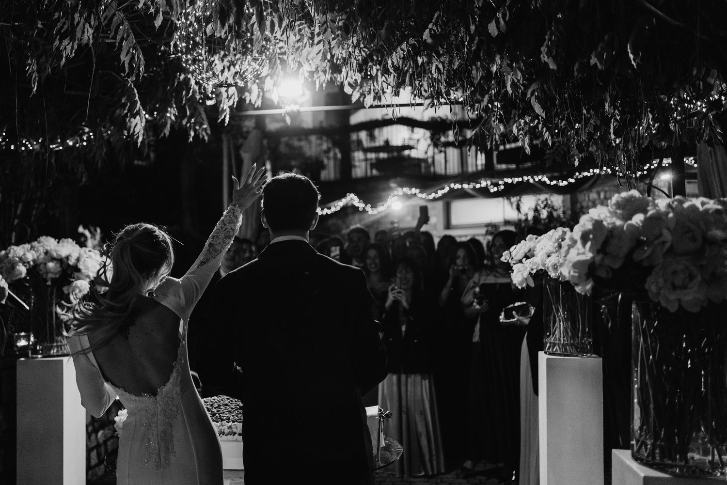 Black and white photo of bride waving to wedding guests among floral arrangements at an outdoor night reception.