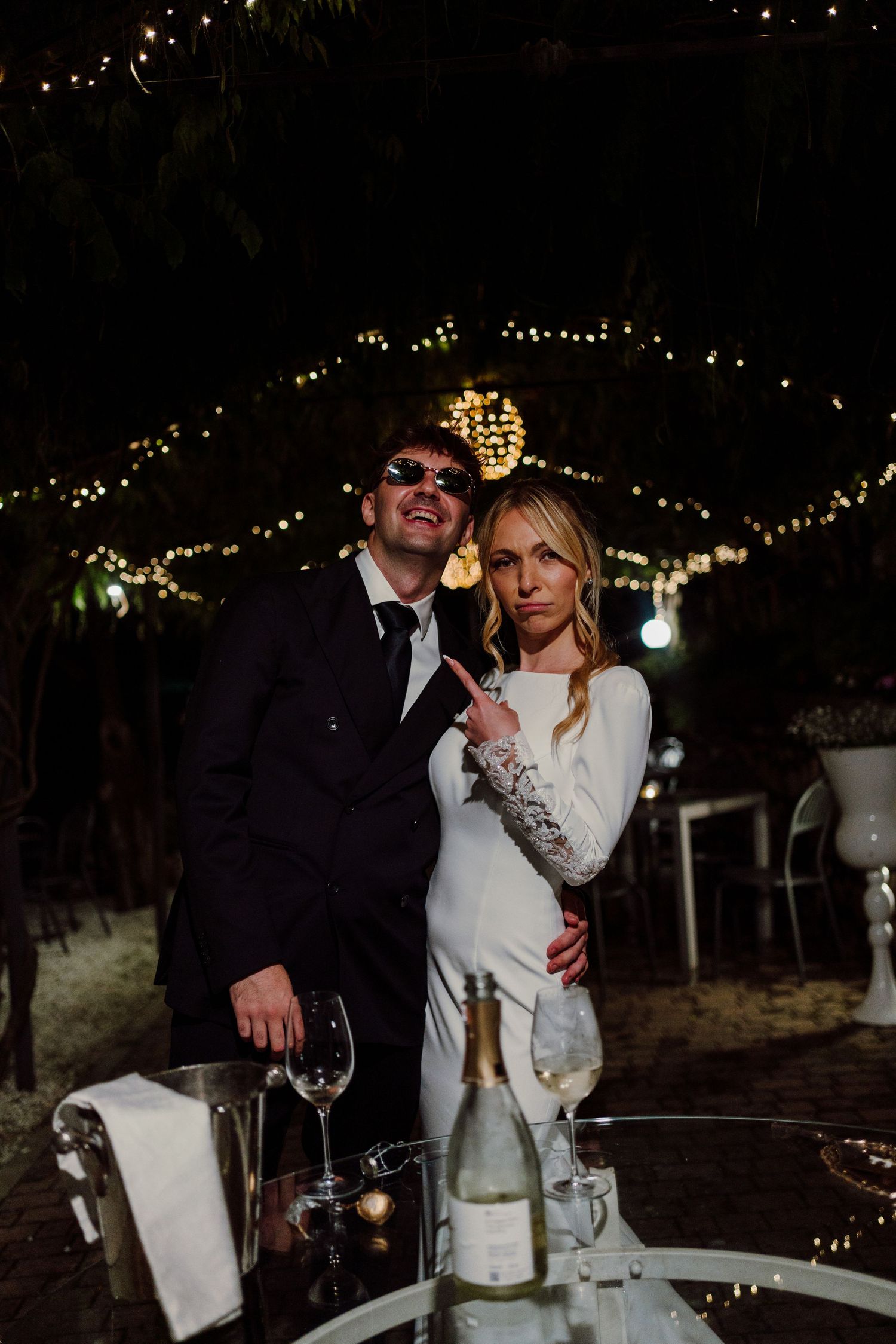 Bride and groom posing playfully with sunglasses and champagne under twinkling fairy lights at night wedding.