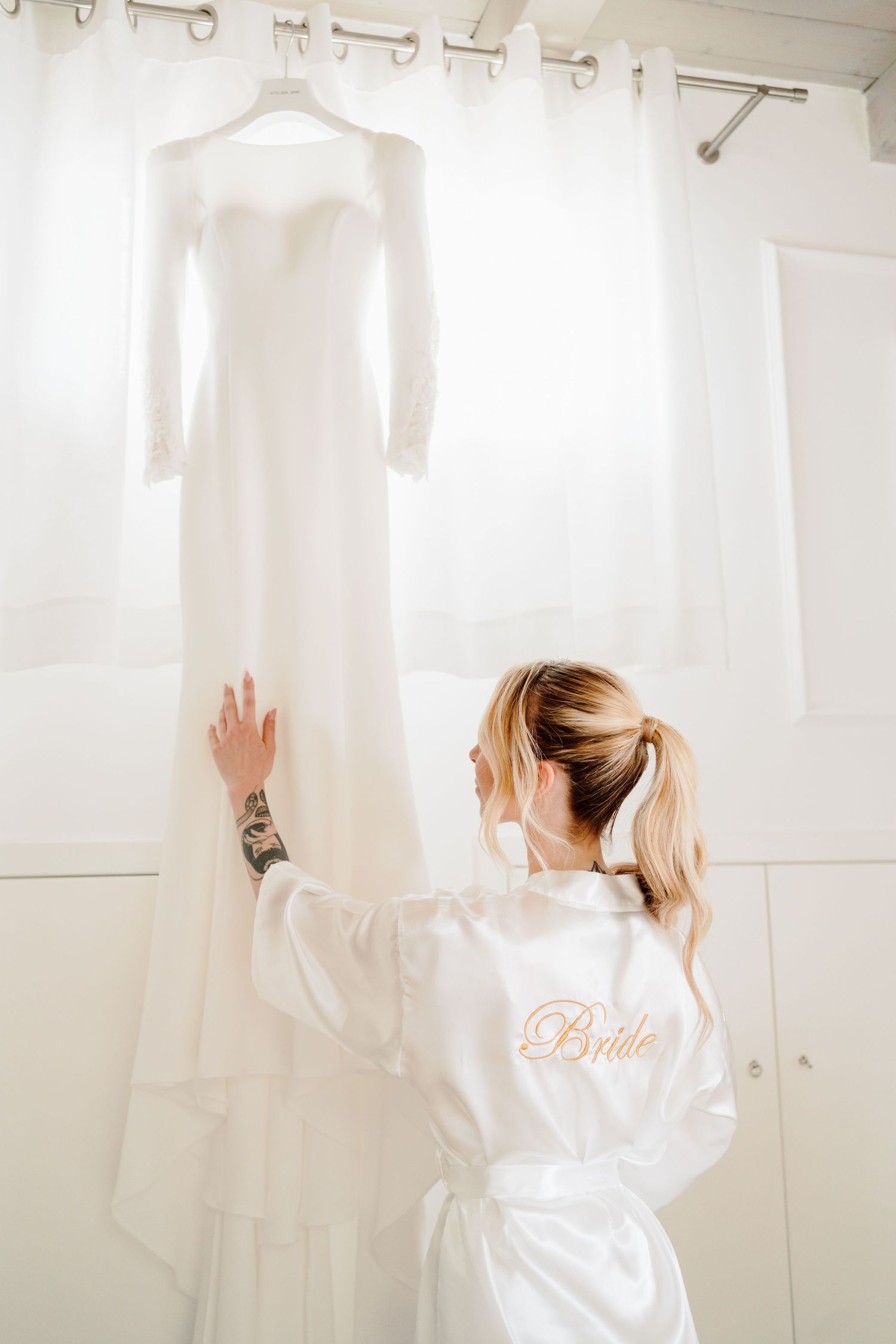 Bride in white robe reaching up to touch her wedding dress hanging in light.