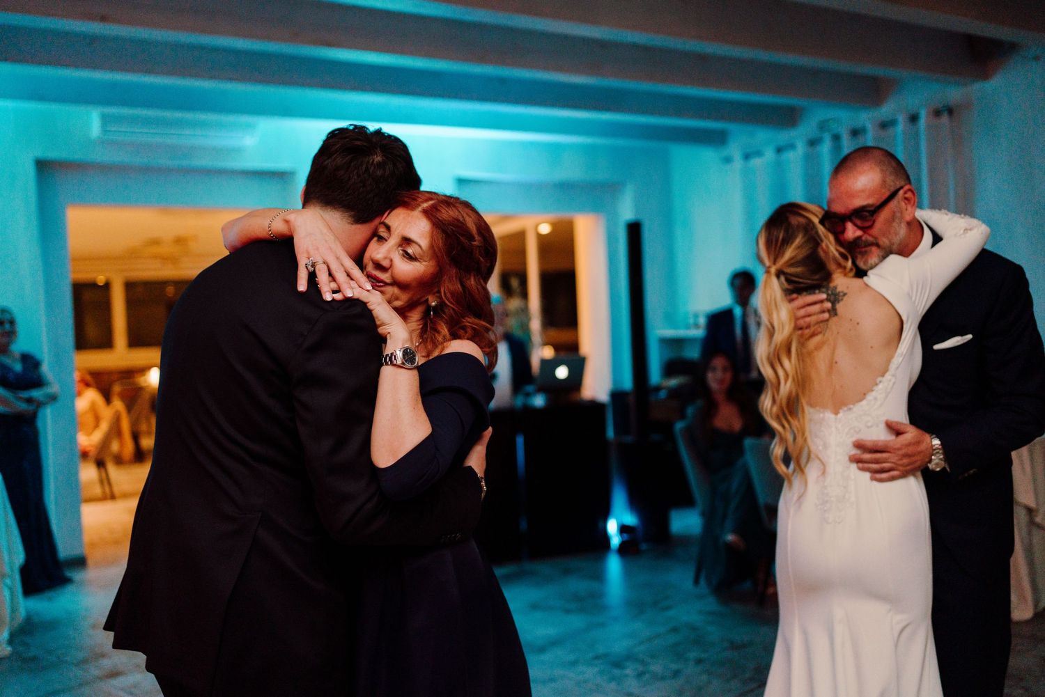 Two couples sharing tender dance floor embraces under blue lighting at a wedding reception.