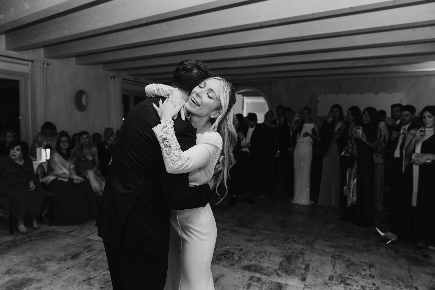 Black and white photo of bride and groom dancing closely surrounded by wedding guests on dance floor.