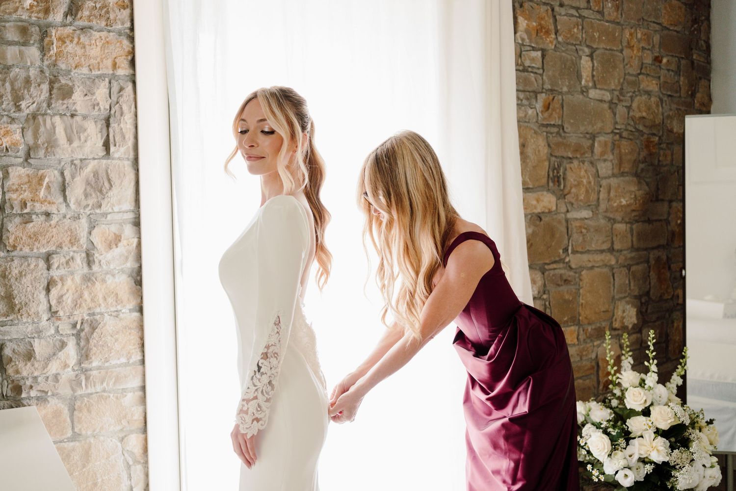 Bridesmaid in burgundy dress helping bride into elegant long-sleeve lace wedding gown.