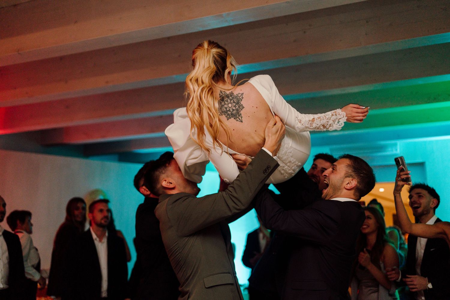 Groomsmen lift smiling bride with back tattoo high in the air at wedding reception.