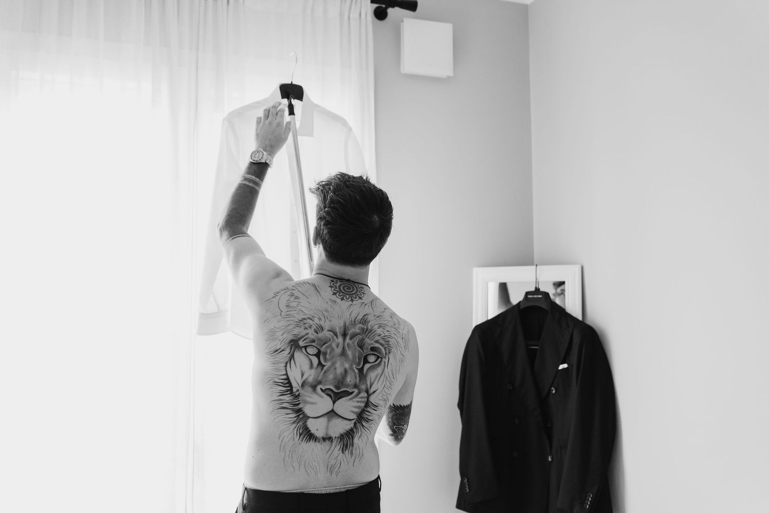 Black and white photo of tattooed groom with lion back tattoo reaching for shirt on hanger.